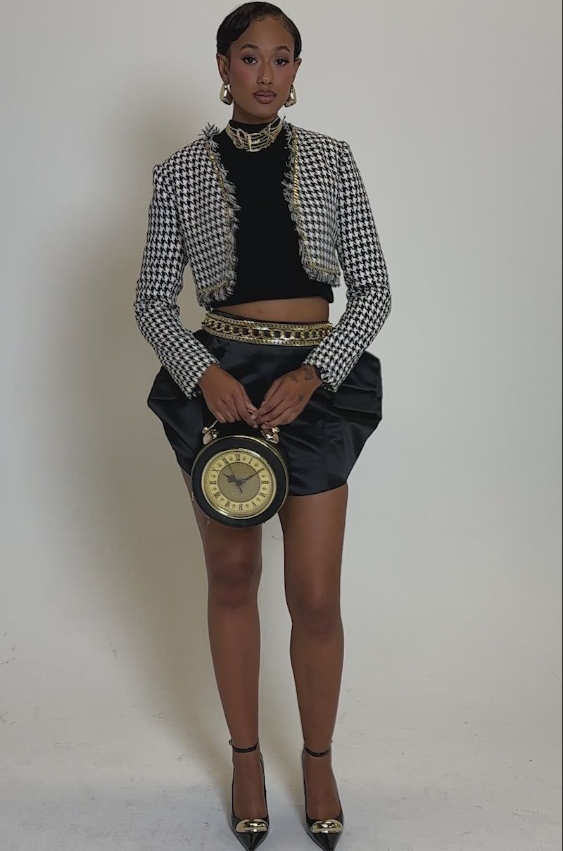 Video of woman modeling the ALL FOR IT MINI SKIRT, a black crop top, houndstooth blazer, black heels with gold accents, and holding a round black handbag with gold details.