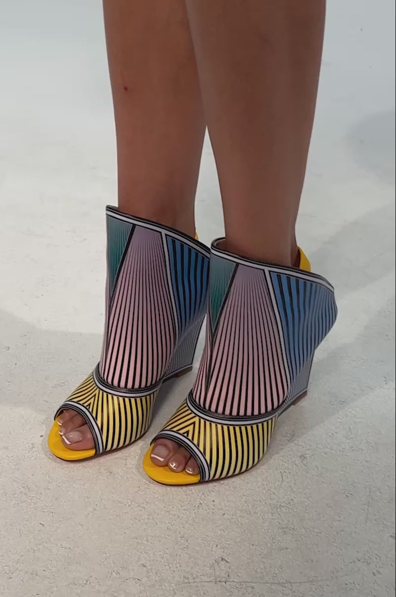 Video of woman modeling the AKIRA GOLD WEDGE OPEN TOE GEOMETRIC BOOTIE, featuring colorful striped patterns in blue, yellow, pink, and green, a sculptural wedge heel, peep toe design, and bold color blocking against a plain background.