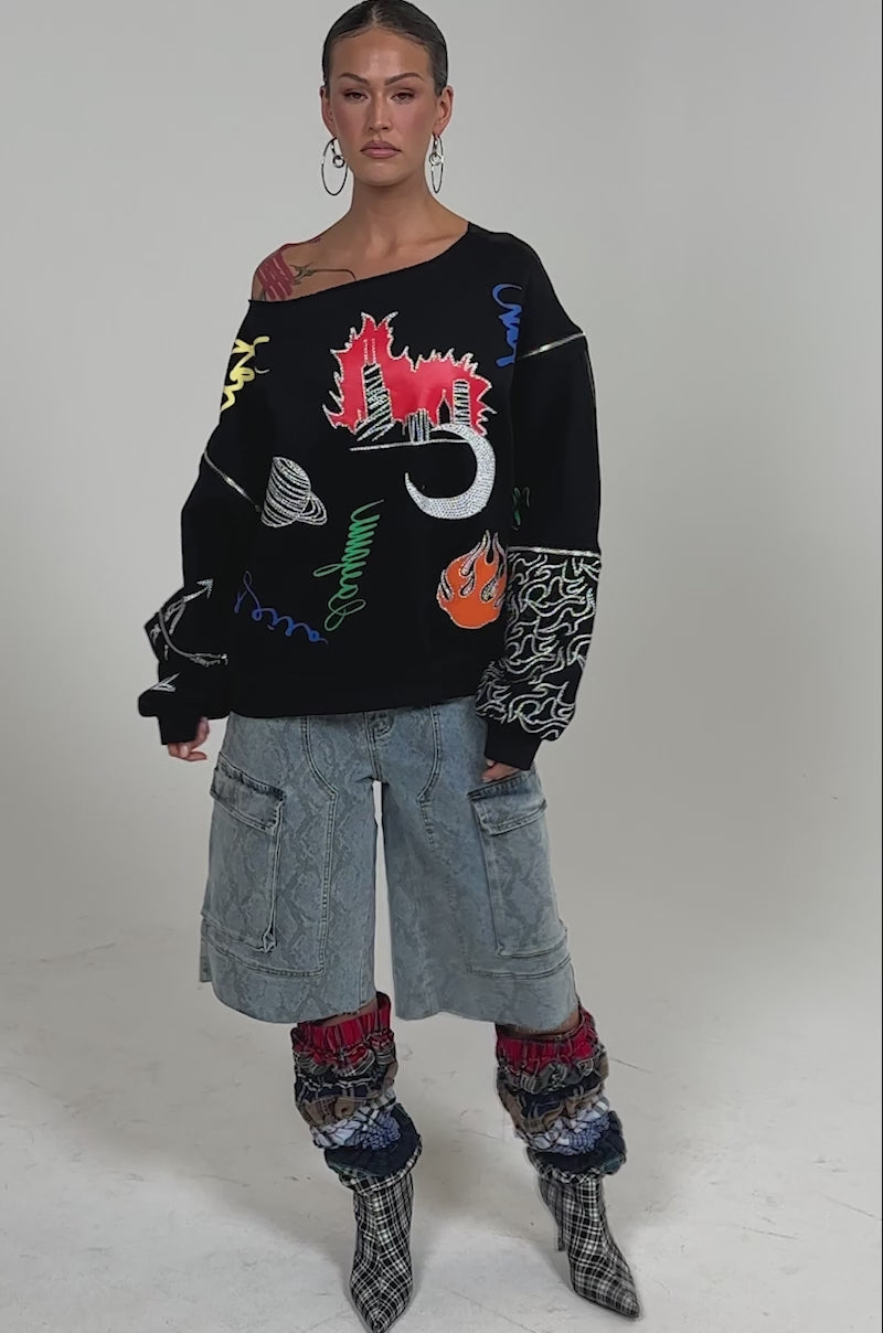 Video of woman modeling the DEEP THOUGHTS OVERSIZED GRAPHIC SWEATSHIRT with colorful rhinestone prints, hoop earrings, and light blue cargo jeans posing against a plain background, showing shoulder tattoos and a neutral expression.