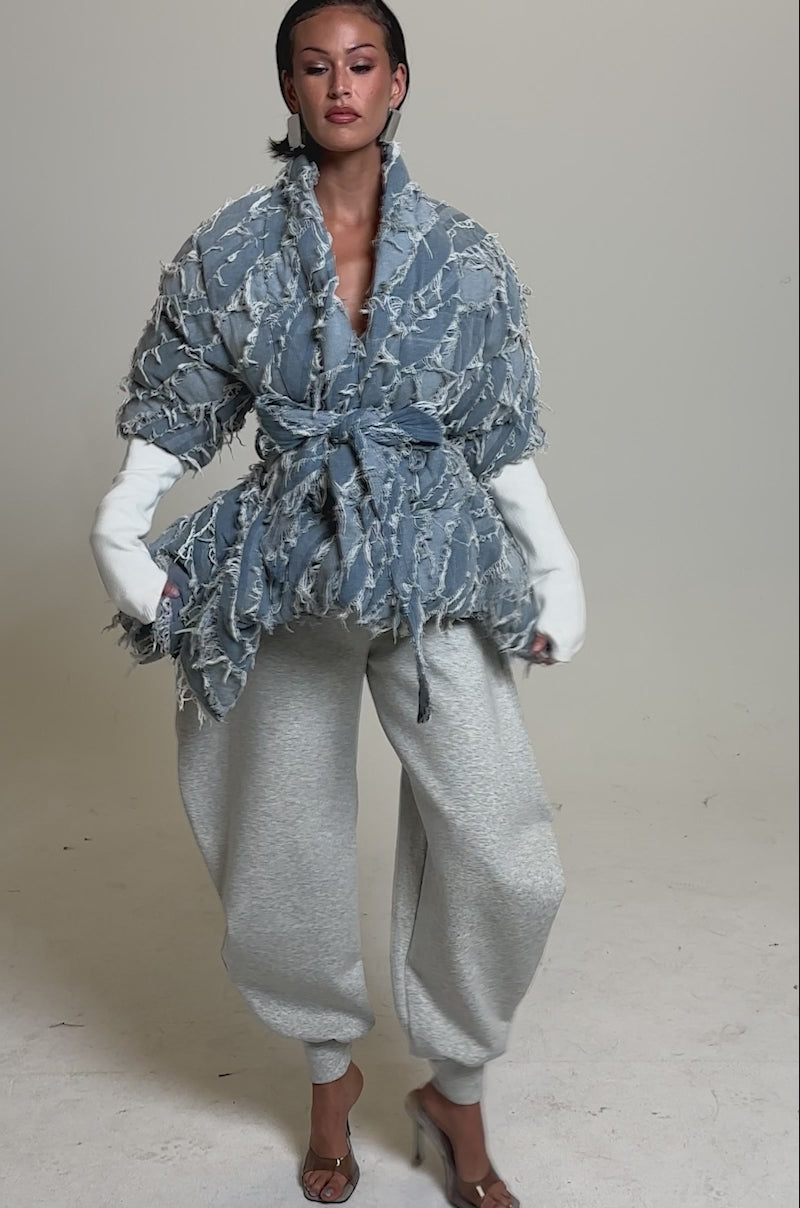 A video of a woman in the RIB SLEEVE PEPLUM PUFFER IN DENIM over a white top, paired with loose gray sweatpants and transparent heels, set against a plain light background.