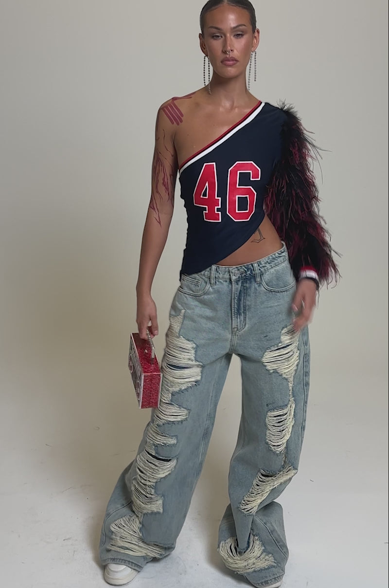 Video of woman modeling the SUPERBOWL PARTY ONE SLEEVE TEE with feathered sleeve and number 46, one hand in her ripped jeans pocket. She carries a small red purse, wears long earrings, and has slicked-back hair.