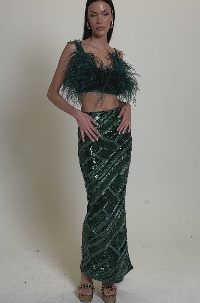 A video of a woman in the FLY AWAY FEATHER BRA TOP and a high-waisted, sequined, geometric-patterned green skirt stands in profile against a plain light gray background.
