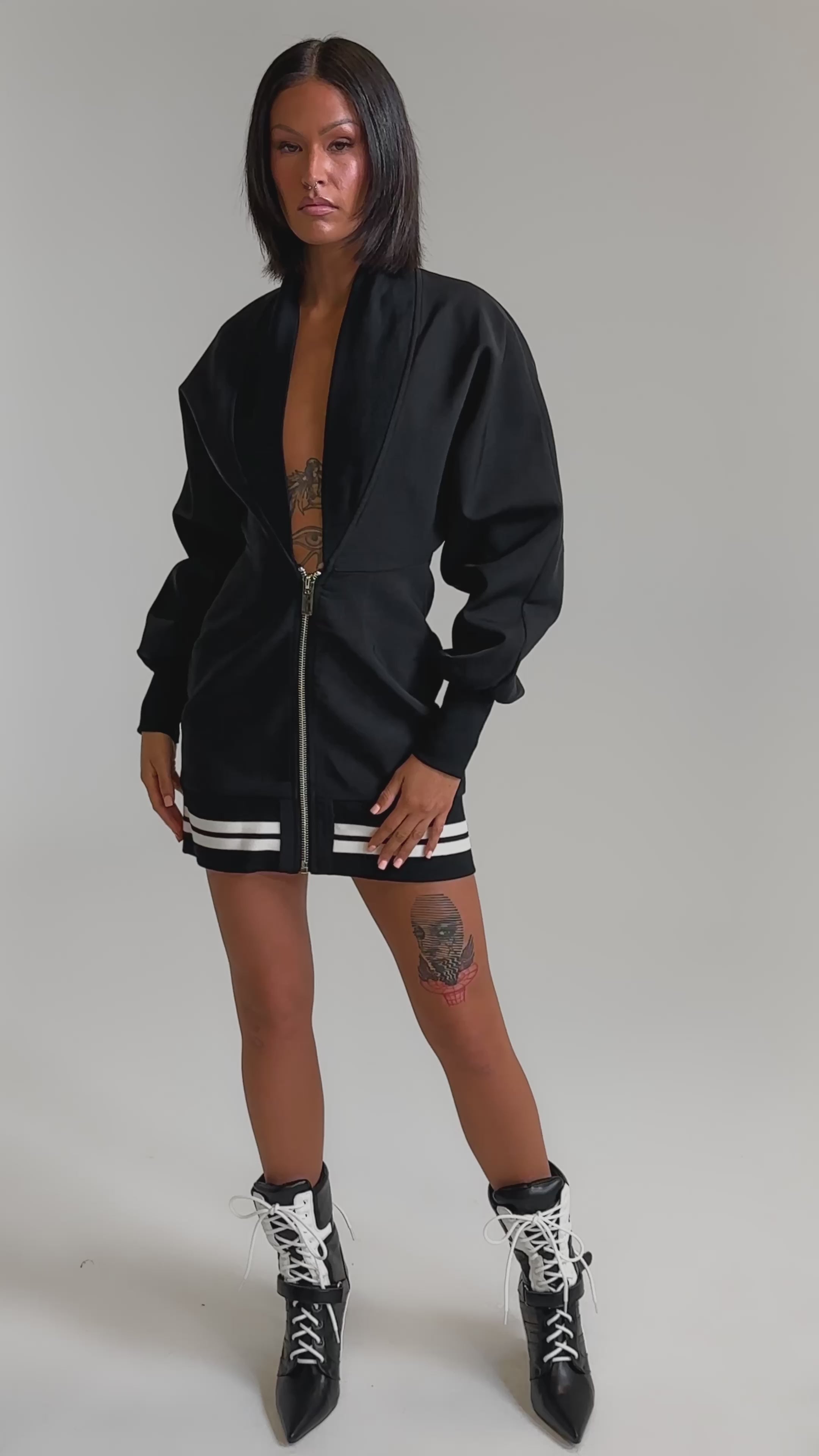 A video of a woman wearing the FAST LANE MINI DRESS, a black zip-up mini dress with white striped hem, paired with black lace-up ankle boots and showing tattoos on her chest and thigh.