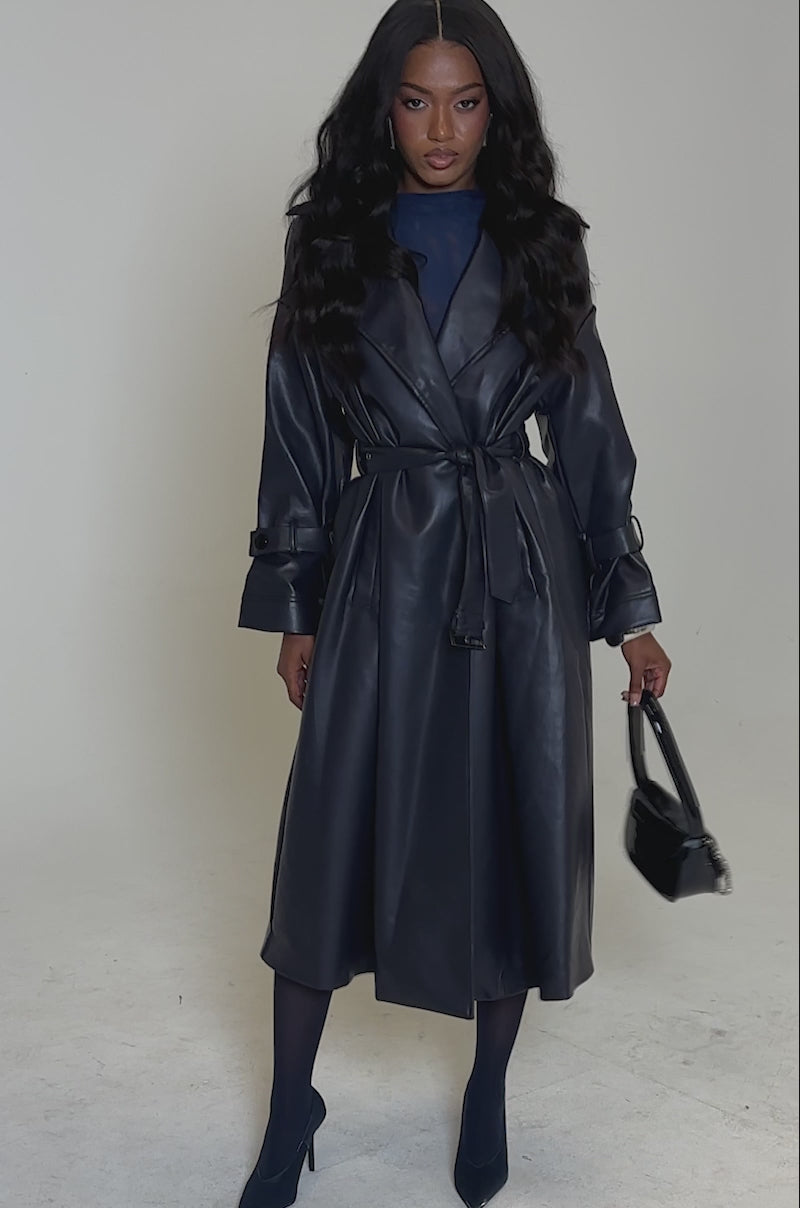 A video of a woman posing against a plain background, wearing the ALINNA FAUX LEATHER TRENCH IN NAVY over a dark top, with matching navy tights and shoes. She has long, wavy hair.