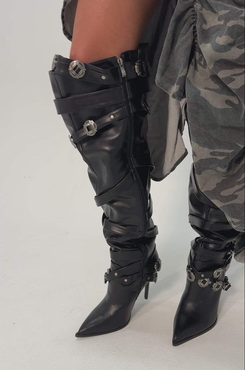 Video of woman modeling the AKIRA GOLD XERXES BLACK STILETTO METAL EMBELLISHED BOOTS, styled with a flowing camouflage-patterned skirt.