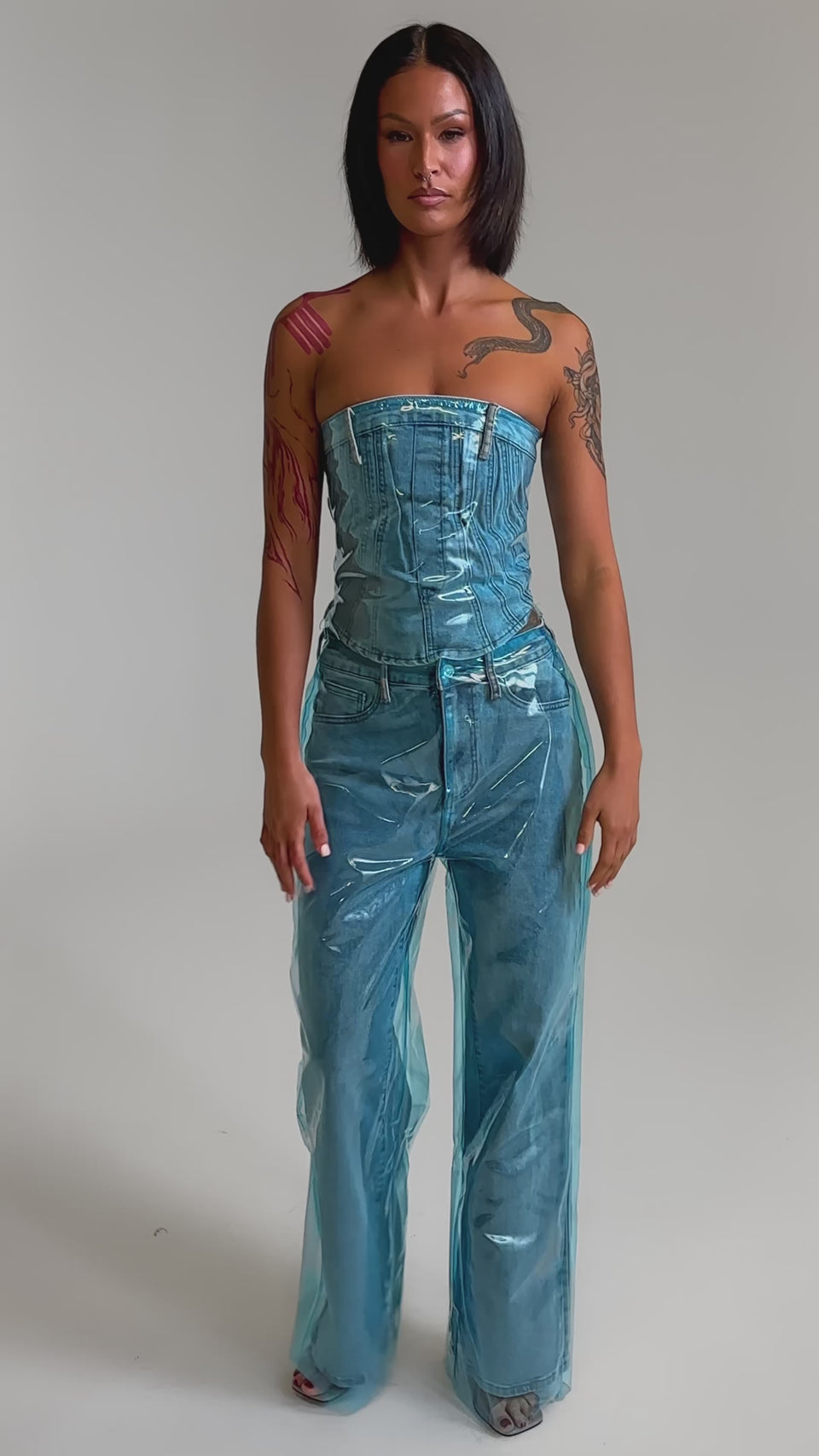 A video of a woman in the ELECTRIC FEEL WIDE LEG DENIM with a strapless clear blue PVC overlay jumpsuit, revealing her arm tattoos against a plain light background.