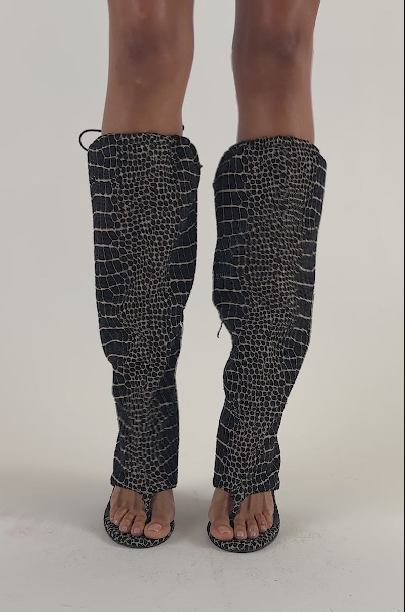 Video of a woman modeling the AZALEA WANG POET BLACK AND WHITE SNAKE PATTERN WEDGE SANDAL, featuring open toes and a knee-high shaft with a black-and-white textured snake pattern that covers their legs up to the knees.