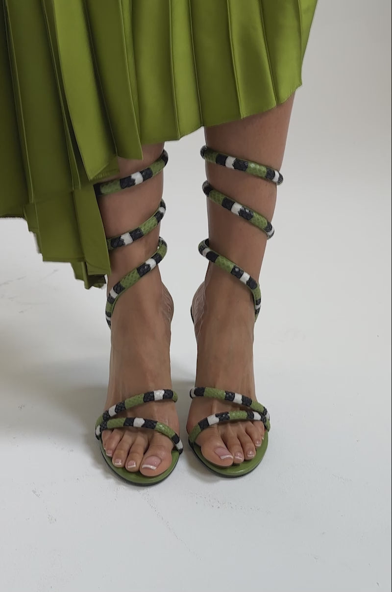 A video of a person in a pleated olive-green skirt and AKIRA GOLD CITLALI GREEN SNAKE COIL SANDALS with snakeskin straps wrapped around their ankles and calves.
