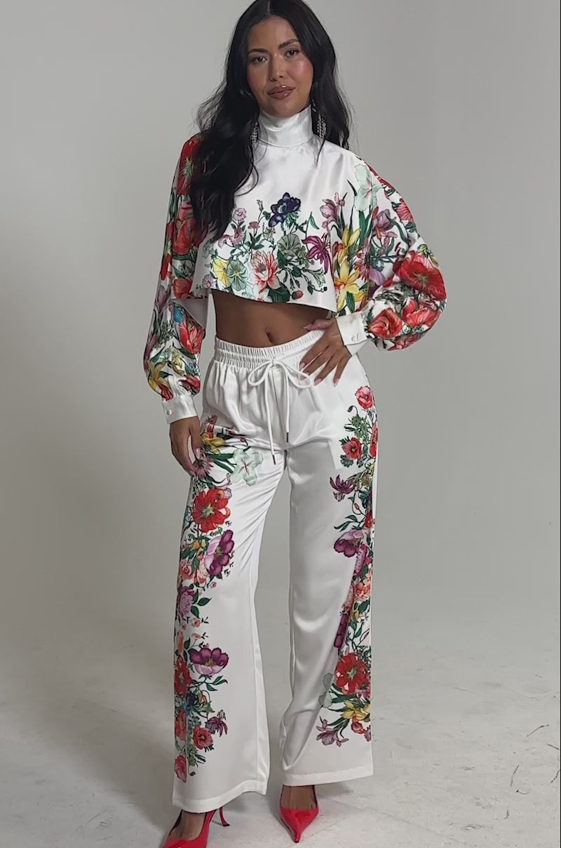 Video of woman modeling the FULL BLOOM STRAIGHT LEG TROUSERS in a floral print, paired with a white, long-sleeve crop top and red pointed heels, creating a fresh spring look against a light background.