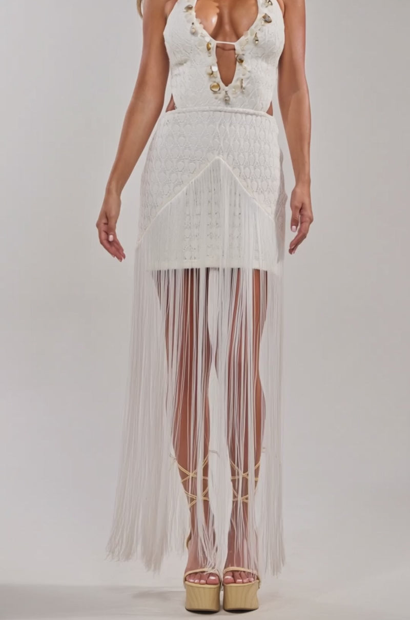 Video of woman modeling the SIREN EYES EMBELLISHED MAXI DRESS, featuring a white hue, deep neckline, gold accents, and long fringe. She pairs it with a textured beige clutch and strappy heels, posing against a plain gray background.