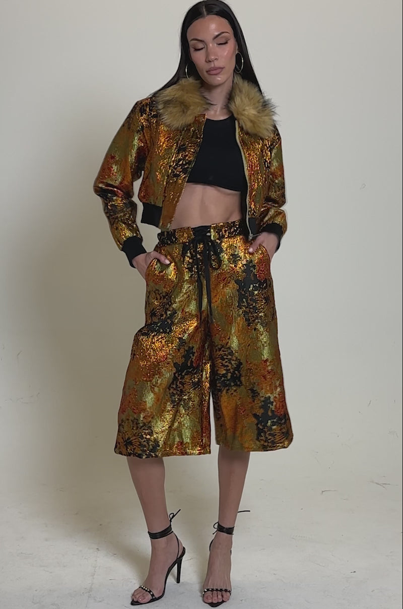 A video of a woman wearing the TWIN FLAME BROCADE BOMBER with a faux fur collar, styled with a black crop top, matching knee-length skirt, and black strappy heels against a simple neutral background.