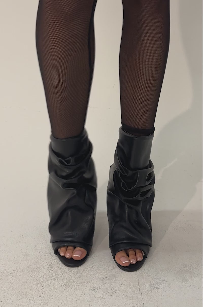 A video of a model wearing a black jacket, sheer black stockings, and AZALEA WANG APEX Black Slouchy Open Toe Booties.