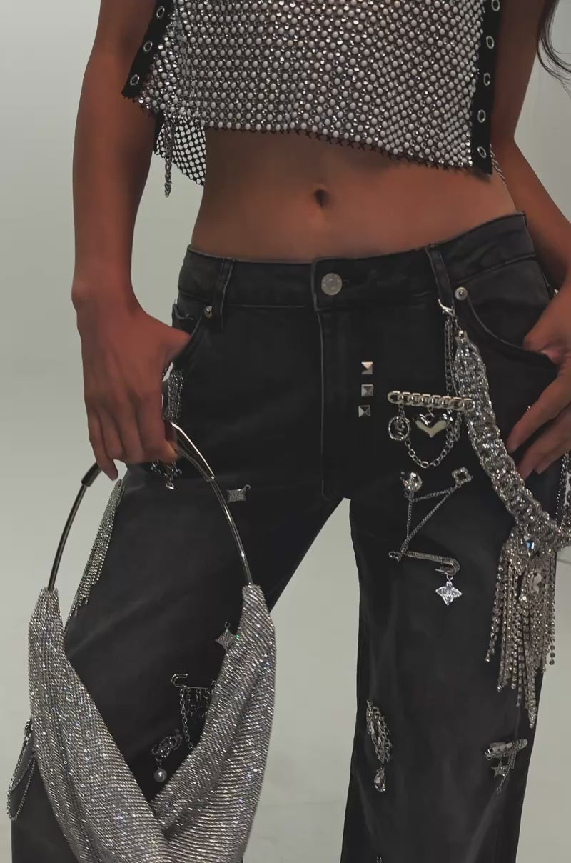 Video of a woman modeling THE BEST ONE YET RELAXED FIT BLING DENIM JEANS, decked out in silver chains, safety pins, and charm embellishments in true streetwear style, holds a textured gray bag. Only the lower torso and legs are visible.
