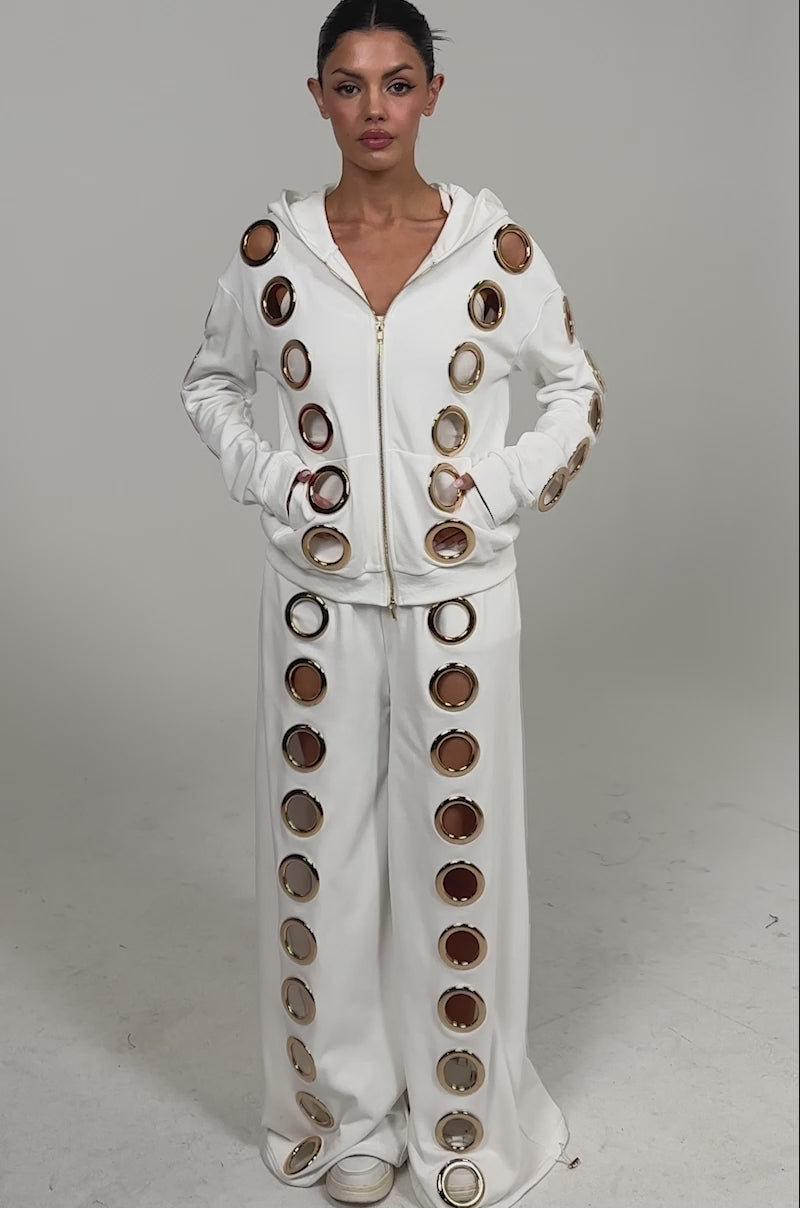Video of woman modeling the THROUGH THE NIGHT WIDE LEG SWEATPANT WITH HOLES IN WHITE, paired with a luxe athleisure white hoodie, both featuring bold gold grommet ring details, against a plain light background.