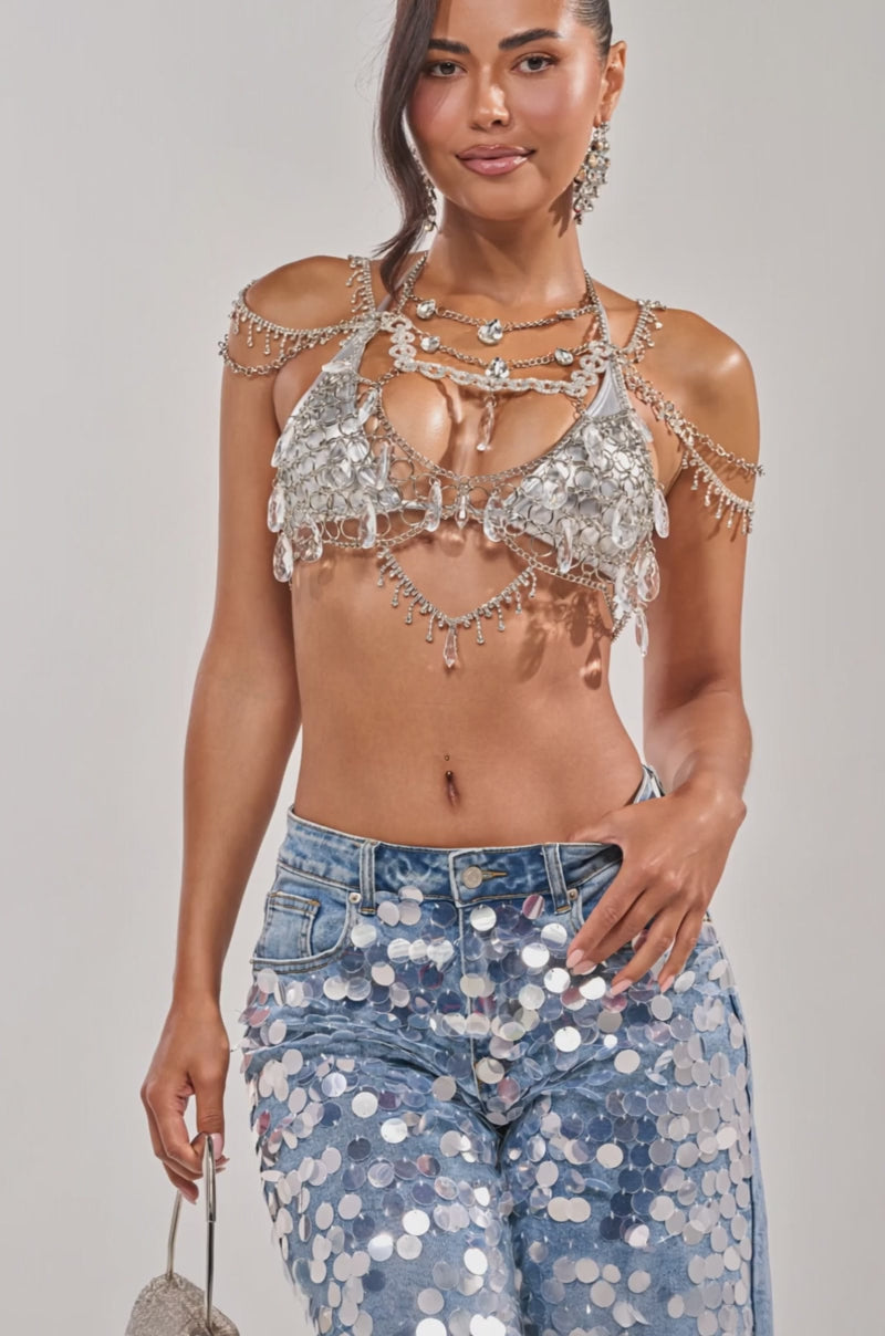 Video of woman modeling the DRIPPY RHINESTONE CHAIN TOP with jeweled straps and silver-sequined light blue jeans. Her long dark hair is pulled back, accessorized with bold earrings, embodying festival-ready confidence and elegance.