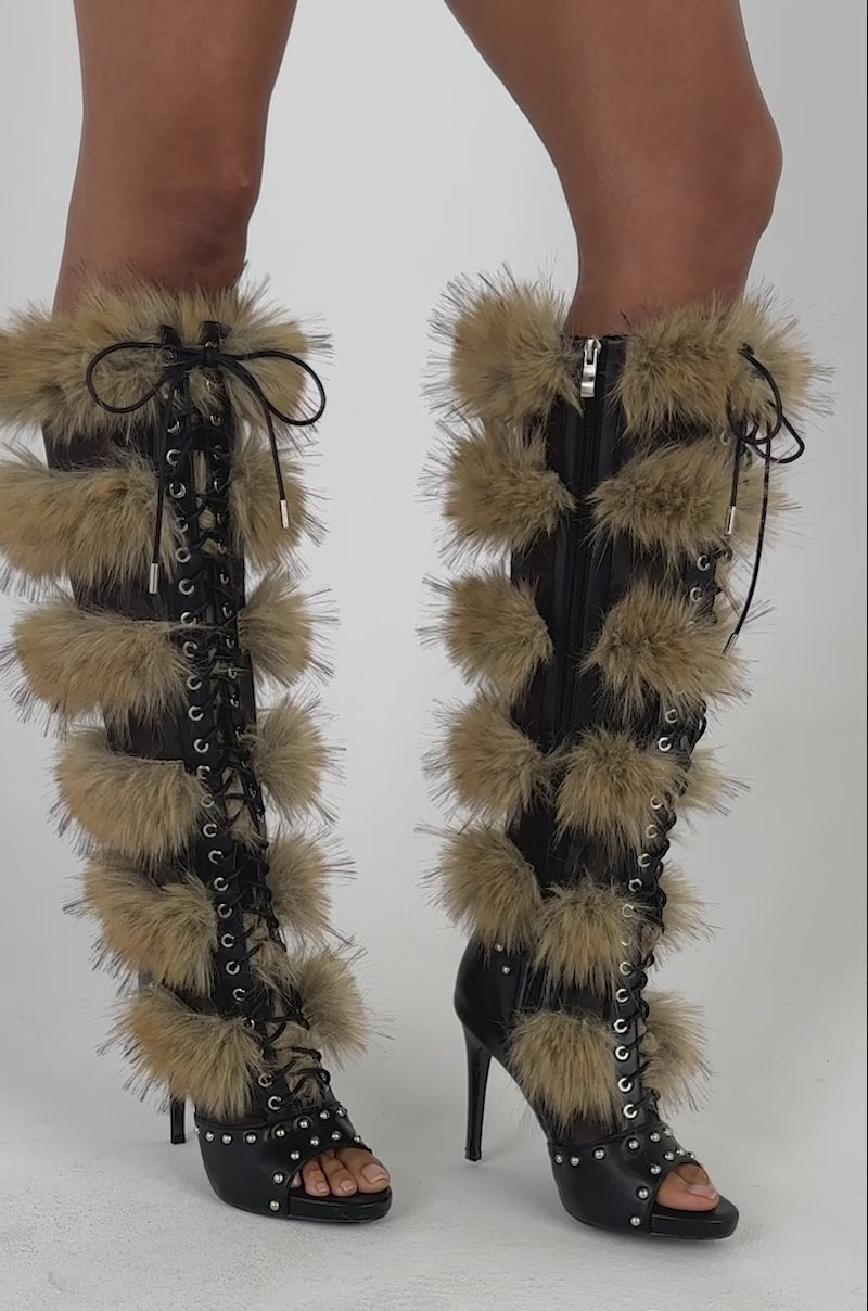 Video of a woman modeling the AZALEA WANG EMBRY TAN MULTI FUR OPEN TOE BOOT, featuring tall black lace-up styling and fluffy brown faux fur accents, stands on a white background.