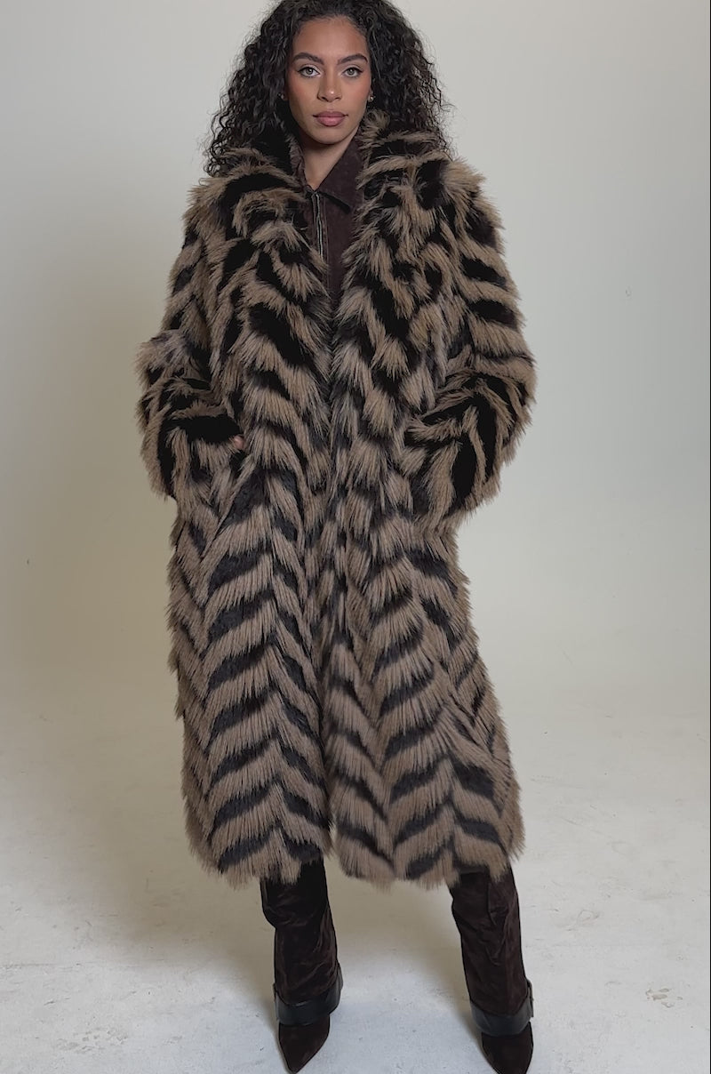 Video of woman modeling the AUTUMN FAUX FUR COAT, a longline coat with a bold brown and black chevron pattern, standing against a plain light background.