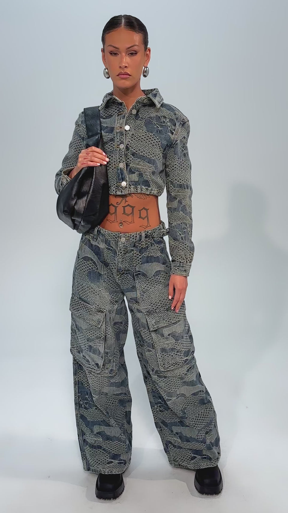 Video of woman modeling the I THINK YOU WANNA KNOW DENIM CARGO PANT, a high-rise, wide-leg style, paired with a blue snakeskin cropped jacket. She carries a black shoulder bag and wears black shoes against a plain light background.
