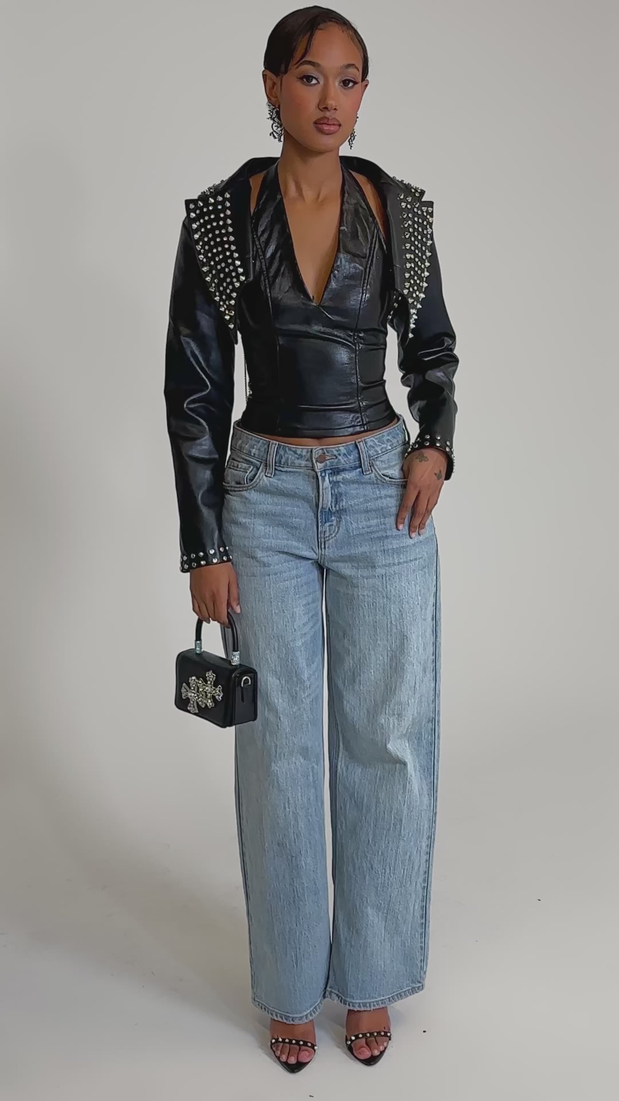 A video of a woman in the NAOMI EMBELLISHED ULTRA CROP MOTO jacket, paired with a black halter top, light blue wide-leg jeans, black heels, a small black handbag, and statement earrings.