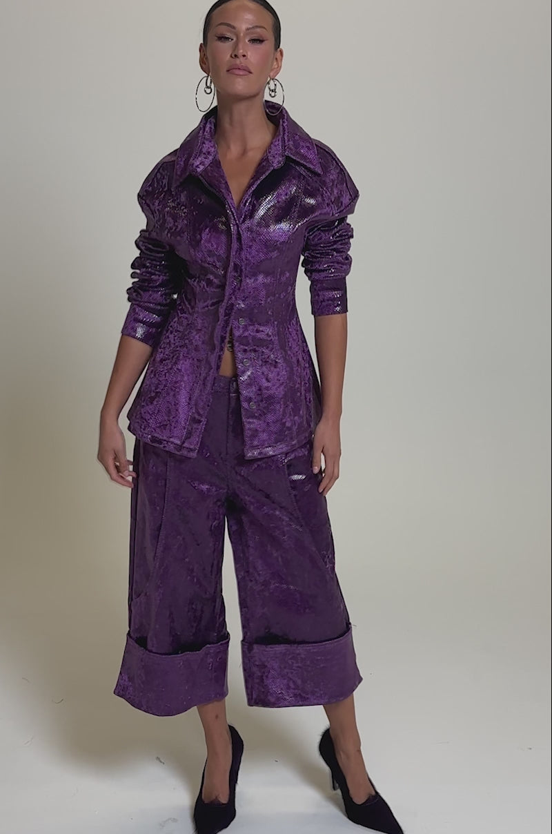 A video of a woman in the shiny purple CRAZY TOUCH VELVET TROUSER suit, featuring wide-leg culottes and a fitted jacket, styled with pointed snakeskin print heels and large hoop earrings against a plain gray background.