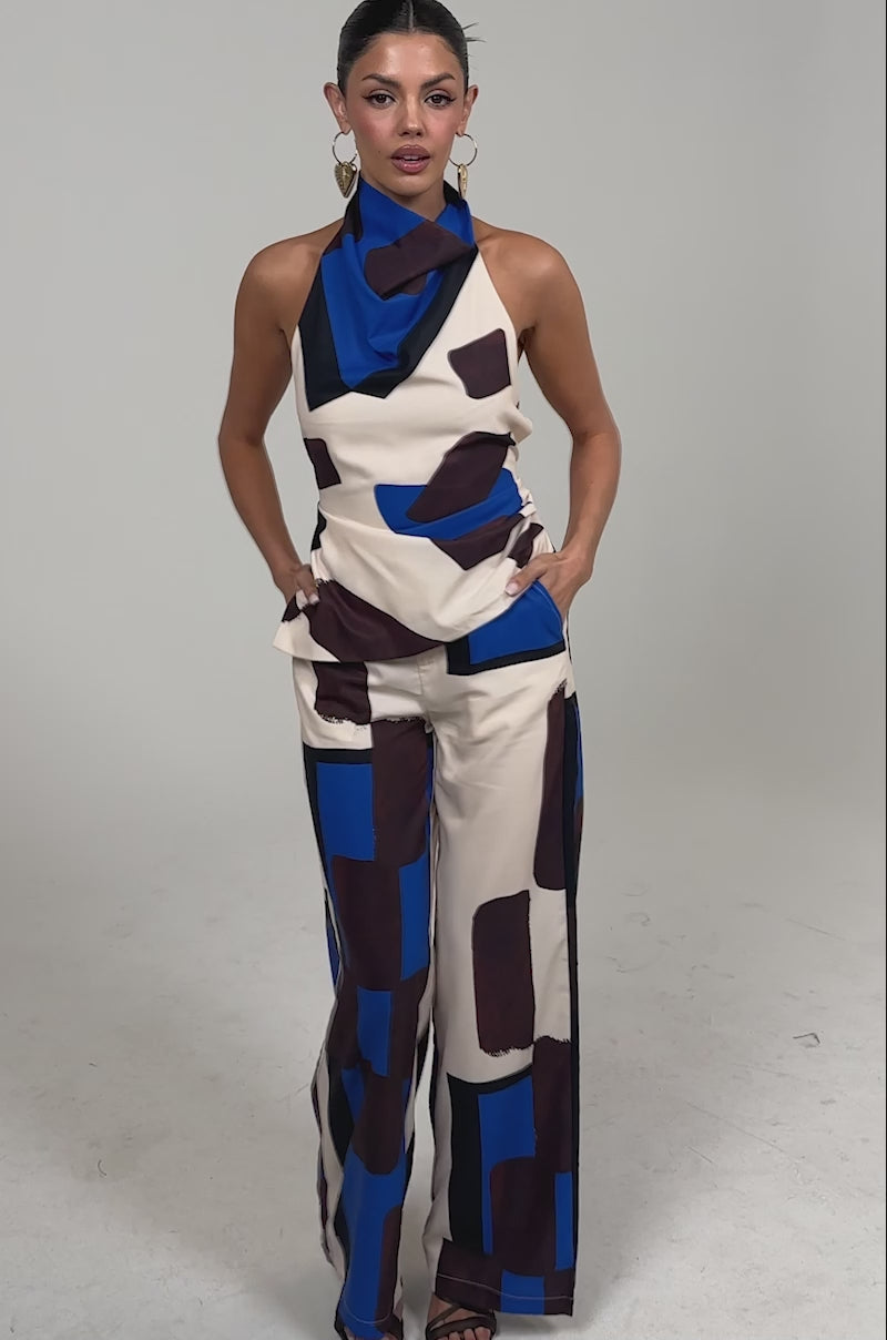 Video of woman modeling the ISLAND TIME TROUSER, a satin wide-leg pant in a bold beige, blue, and burgundy abstract print, paired with a matching halter-neck top, gold earrings, and black heels against a plain background.