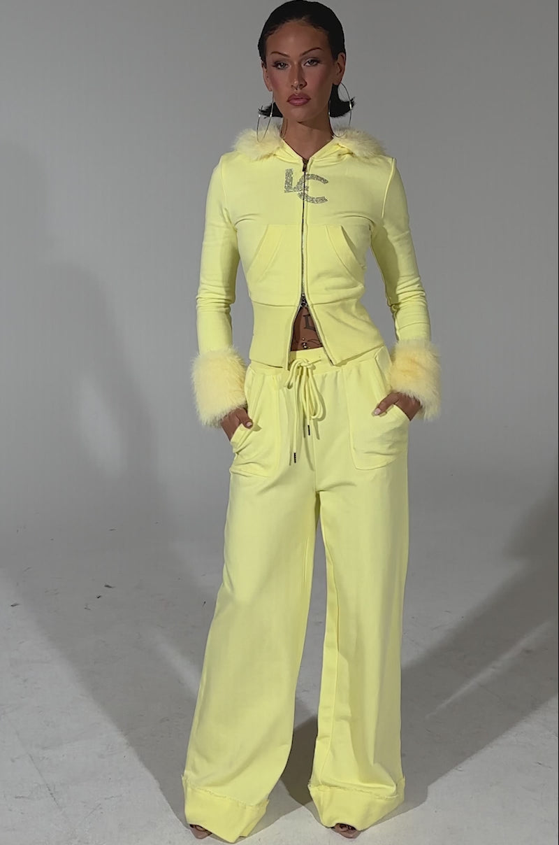 A video of a woman wearing the BUTTERFINGER ZIP UP SWEATSHIRT, a pale yellow velour tracksuit with faux fur hood and cuff trim, and matching wide-leg pants. She poses against a simple, light background.