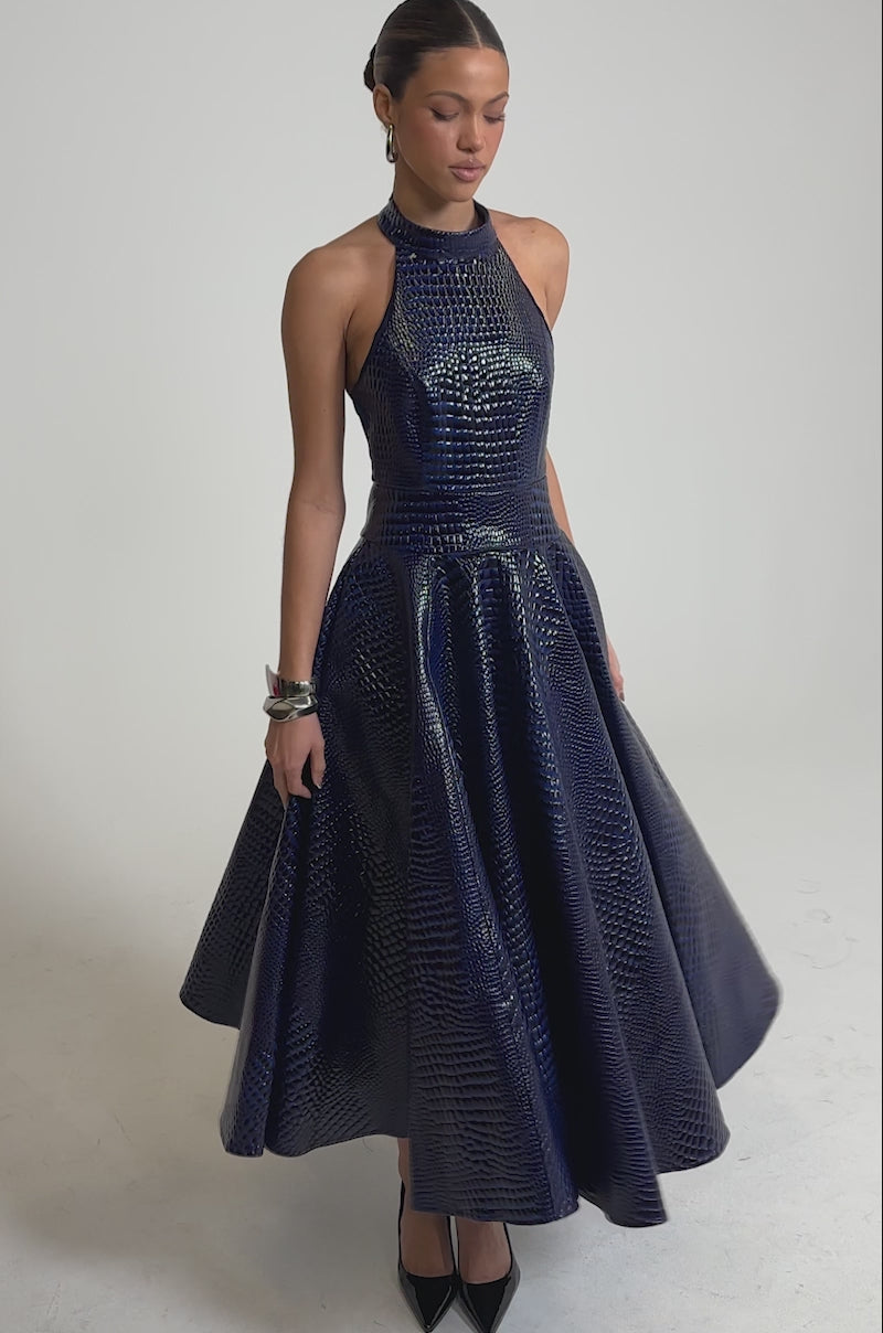 A video of a woman wearing the VEE FAUX LEATHER CROC MIDI DRESS, a sleeveless mock neck royal blue faux leather dress with a croc-textured shine, paired with black pointed heels against a plain light background.