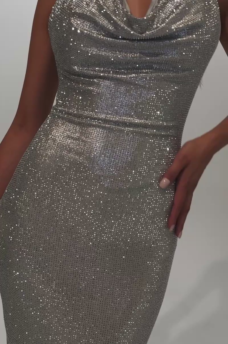 A video of a woman wearing the POPPIN' BOTTLES RHINESTONE MAXI DRESS IN SILVER—a sparkly silver maxi dress with a cowl neck halter neckline—she pairs it with statement earrings, a small clutch, and fringe-accented silver high-heeled sandals.