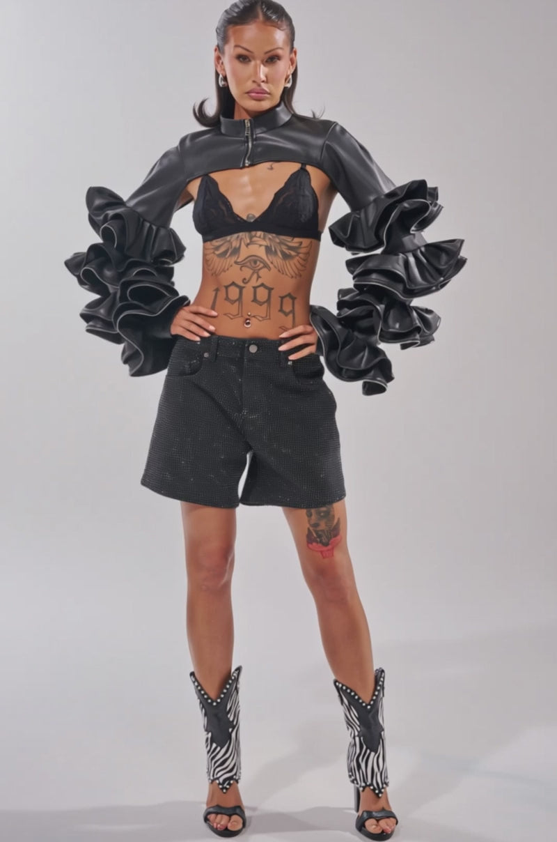 Video of woman modeling the DON'T MESS RUFFLE BOLERO—a cropped black jacket with dramatic ruffled sleeves—paired with a black lace bra and sparkly black shorts.