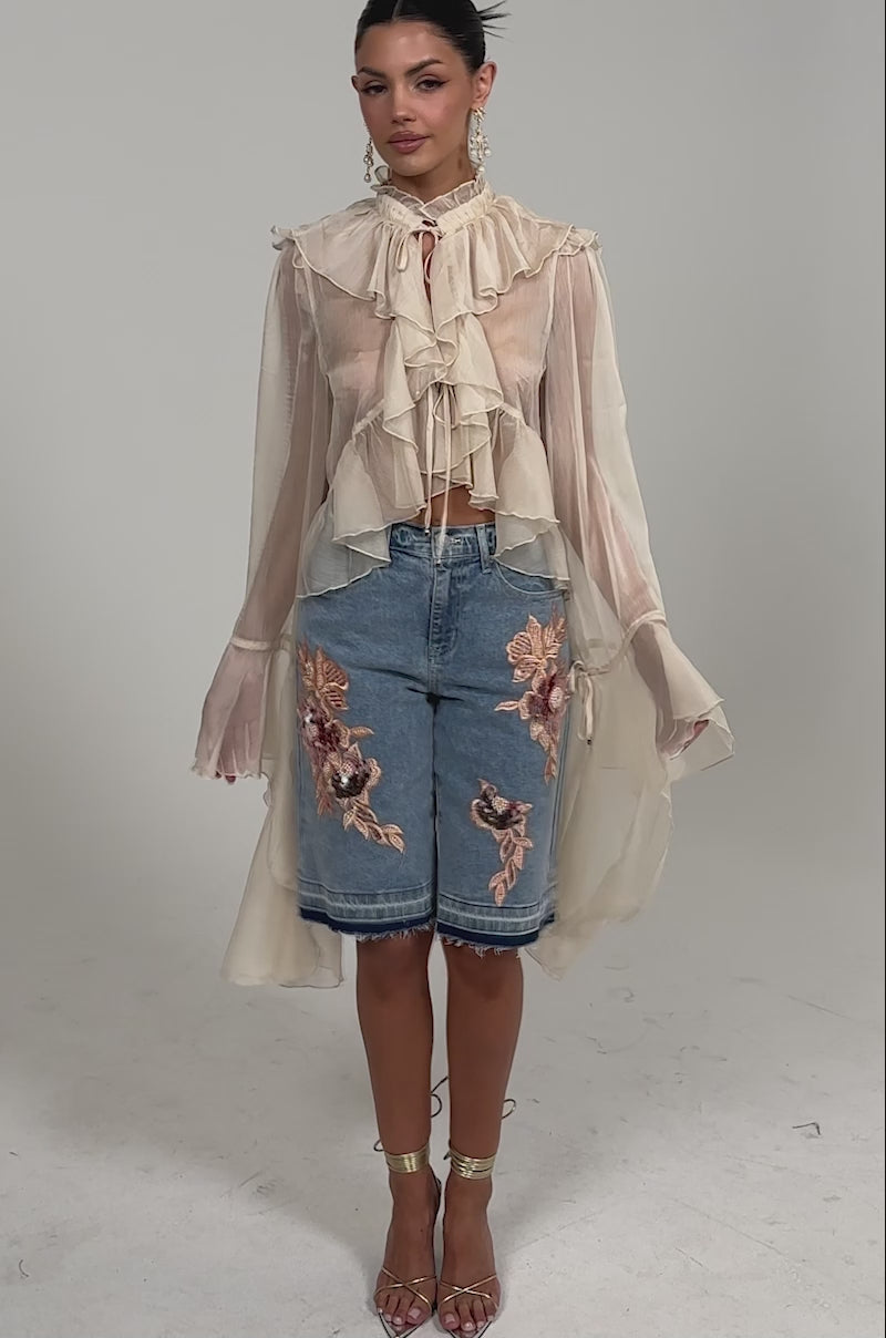 Video of woman modeling the DANCE THE NIGHT AWAY BLOUSE, a sheer ruffle top with dramatic bell sleeves, paired with blue knee-length denim shorts featuring pink floral embroidery, set against a plain white background.