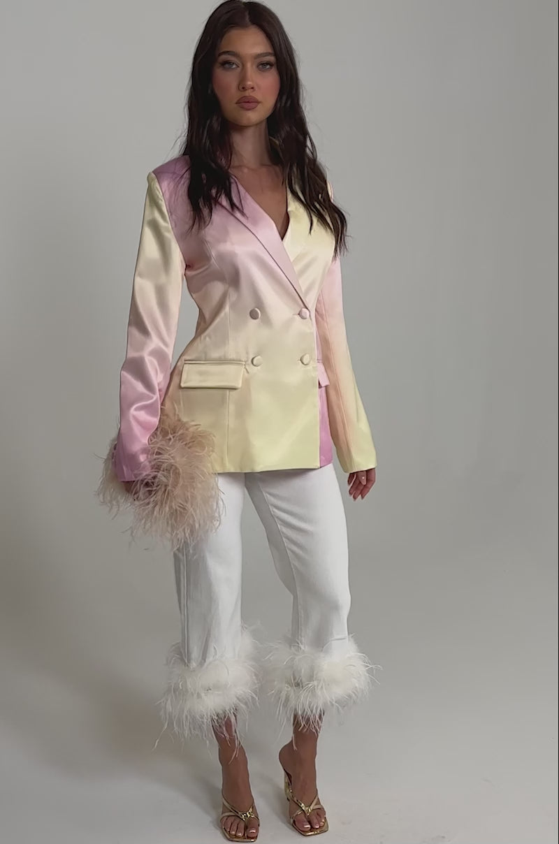 A video of a woman wearing the SWEET TALK SATIN BLAZER in blush pink with double-breasted buttons and feather trim on one sleeve, paired with white cropped pants featuring feathered hems and gold heeled sandals.