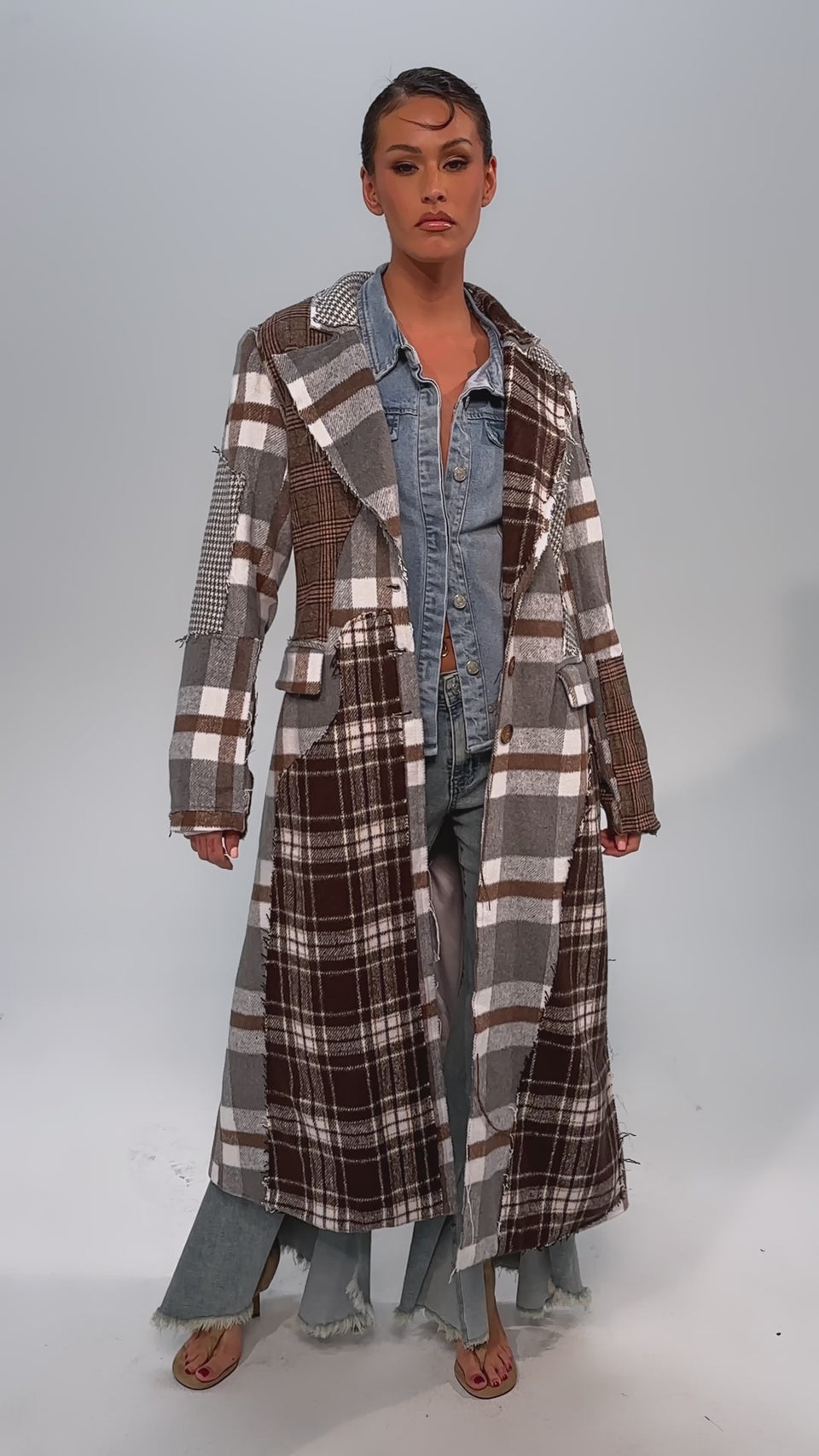 Video of woman modeling the MIXED FEELINGS PATCHWORK TRENCH over a denim shirt and distressed jeans, accessorized with a beige clutch, hoop earrings, and heeled sandals against a plain light gray background.