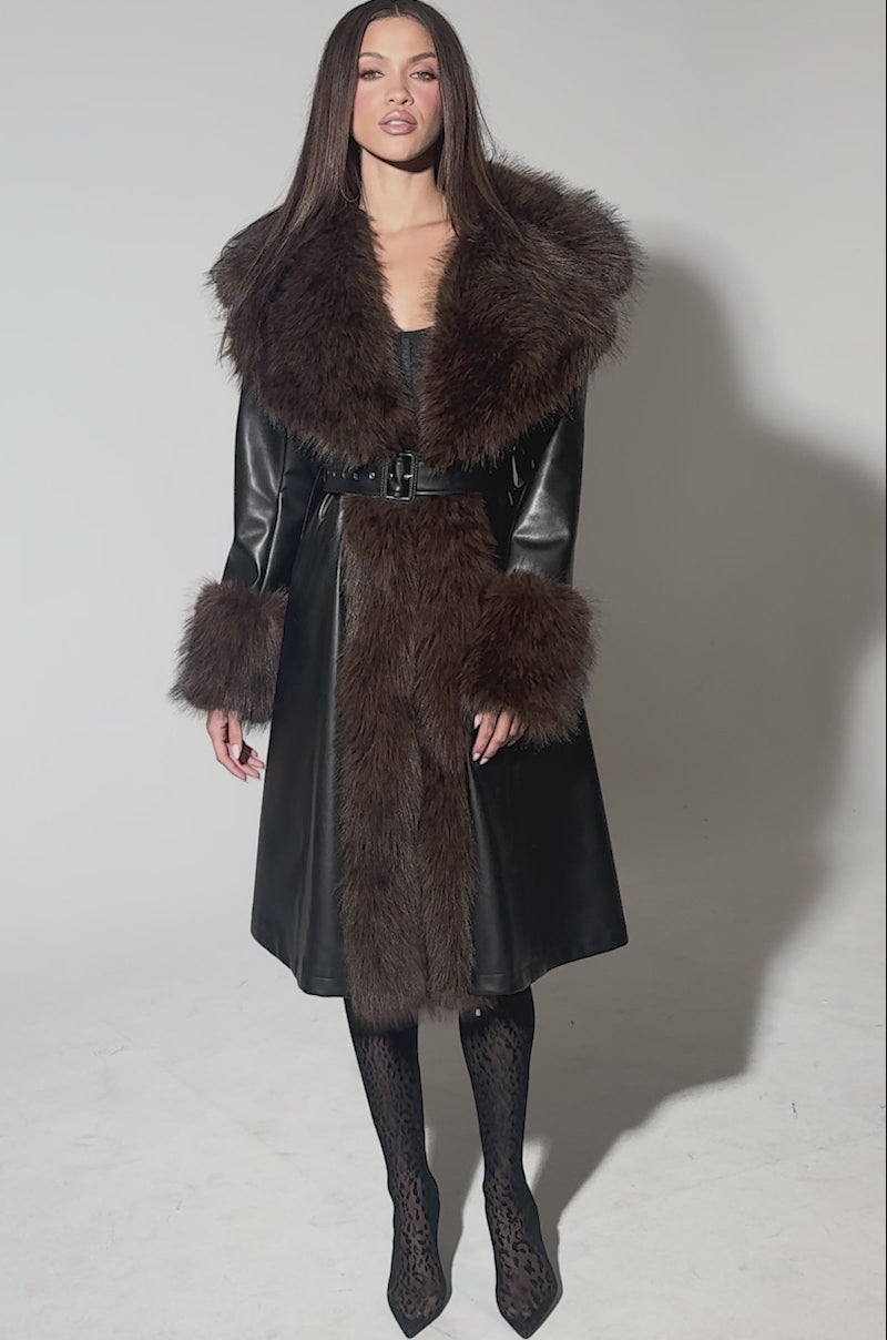 Video of woman modeling the MAIN SQUEEZE FUR TRIM TRENCH, a long black leather coat with a wide brown faux fur collar, matching cuffs, and a belted waist