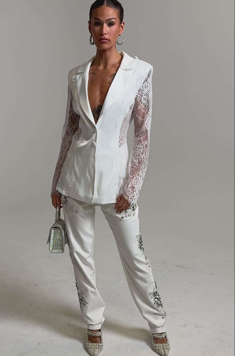 Video of woman modeling the AU CONTRAIRE LACE SATIN BLAZER IN WHITE, featuring sheer lace sleeves. She accessorizes with a silver glittery handbag and hoop earrings, her sleek hair pulled back and a chest tattoo visible.