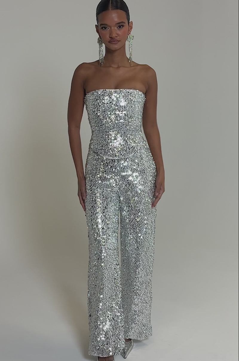 Video of woman modeling the NYE GIRLY TROUSER—a strapless silver sequin corset with high-rise wide-leg pants—styled with pointed silver heels and statement earrings. Her hair is sleek and pulled back.