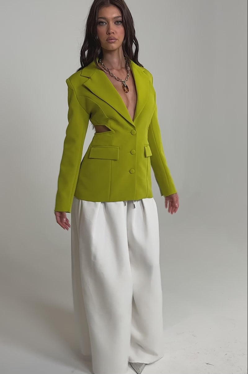 Video of woman modeling the PARTY IN THE BACK CUTOUT BLAZER IN CHARTREUSE with button details and flap pockets, styled over a white skirt and chunky chain necklace, posing against a plain light background.