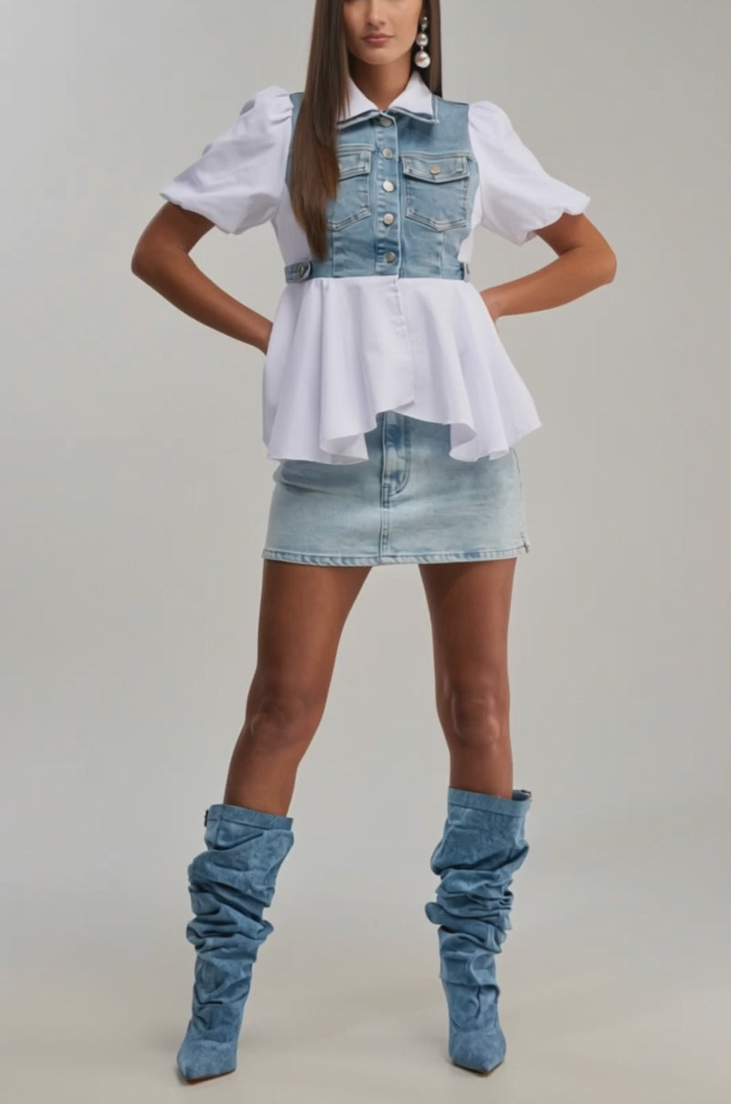 A video of a woman poses in the HOPELESS ROMANTIC DENIM BLOUSE, styled with a light denim mini skirt and slouchy boots, standing against a simple, light background.