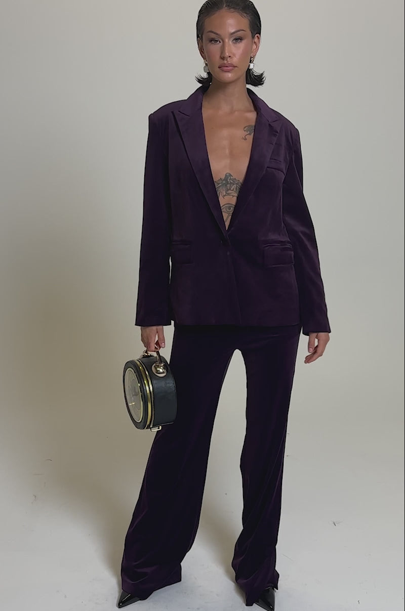 A video of a model wearing the I ADORE YOU BLAZER in deep purple velvet holding a round clock like a handbag against a plain light gray background.