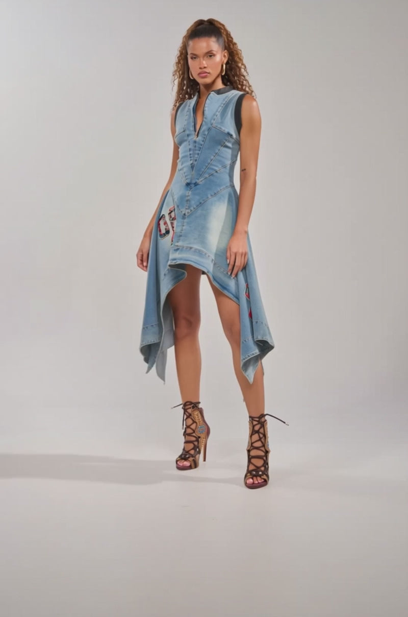 Video of woman modeling the BALLER OFF DUTY DENIM PATCHWORK MINI DRESS, a sleeveless mini featuring an asymmetrical hem, patchwork details, and embroidery. She pairs it with brown lace-up heels against a plain light background.