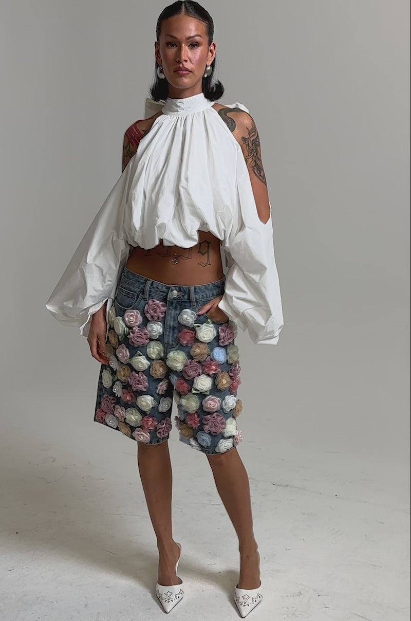 A video of a woman in the IRIS FLORAL BERMUDA SHORT, featuring high-rise denim and 3D floral appliqués, paired with a flowing white blouse with voluminous sleeves. 