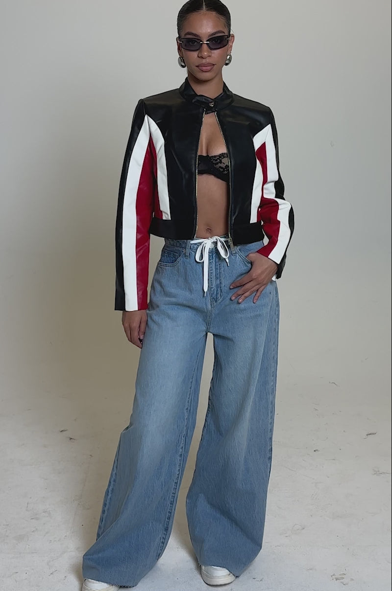 A video of a person wearing sunglasses, the ON YOUR MARKS RACER MOTO cropped black faux leather jacket with white and red stripes, a black bra top, wide-leg light blue drawstring jeans, and white sneakers standing against a plain background.
