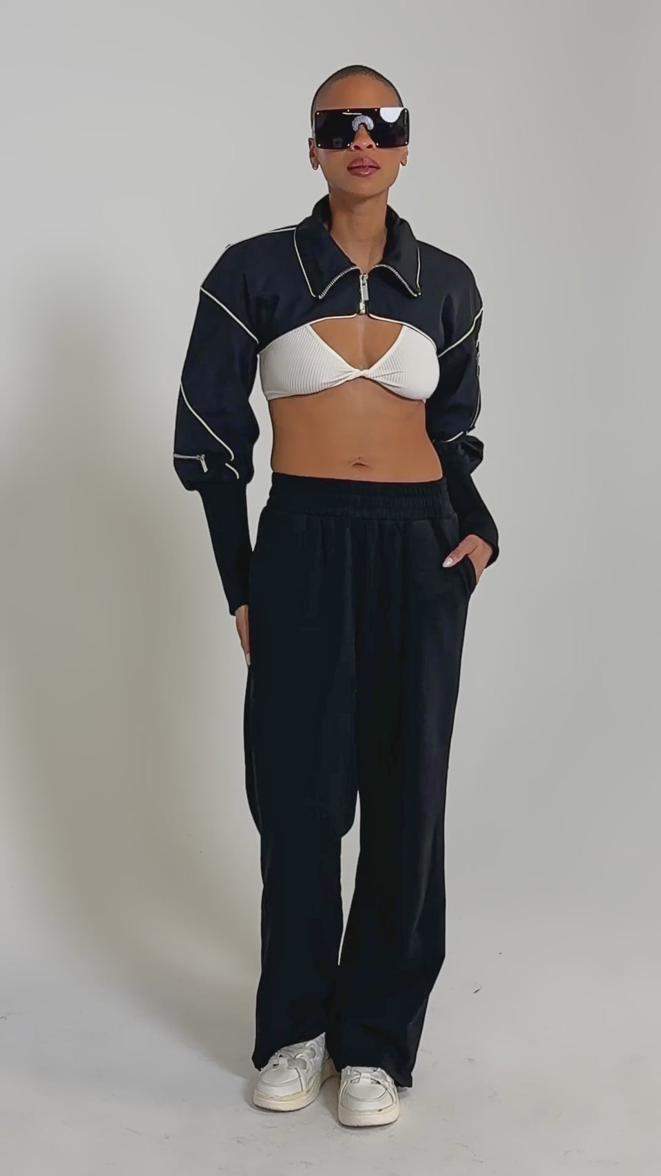 Video of woman modeling the NOW YOU SEE ME SPORTY BOLERO over a white ribbed bralette, and black high-waisted pants against a plain background.
