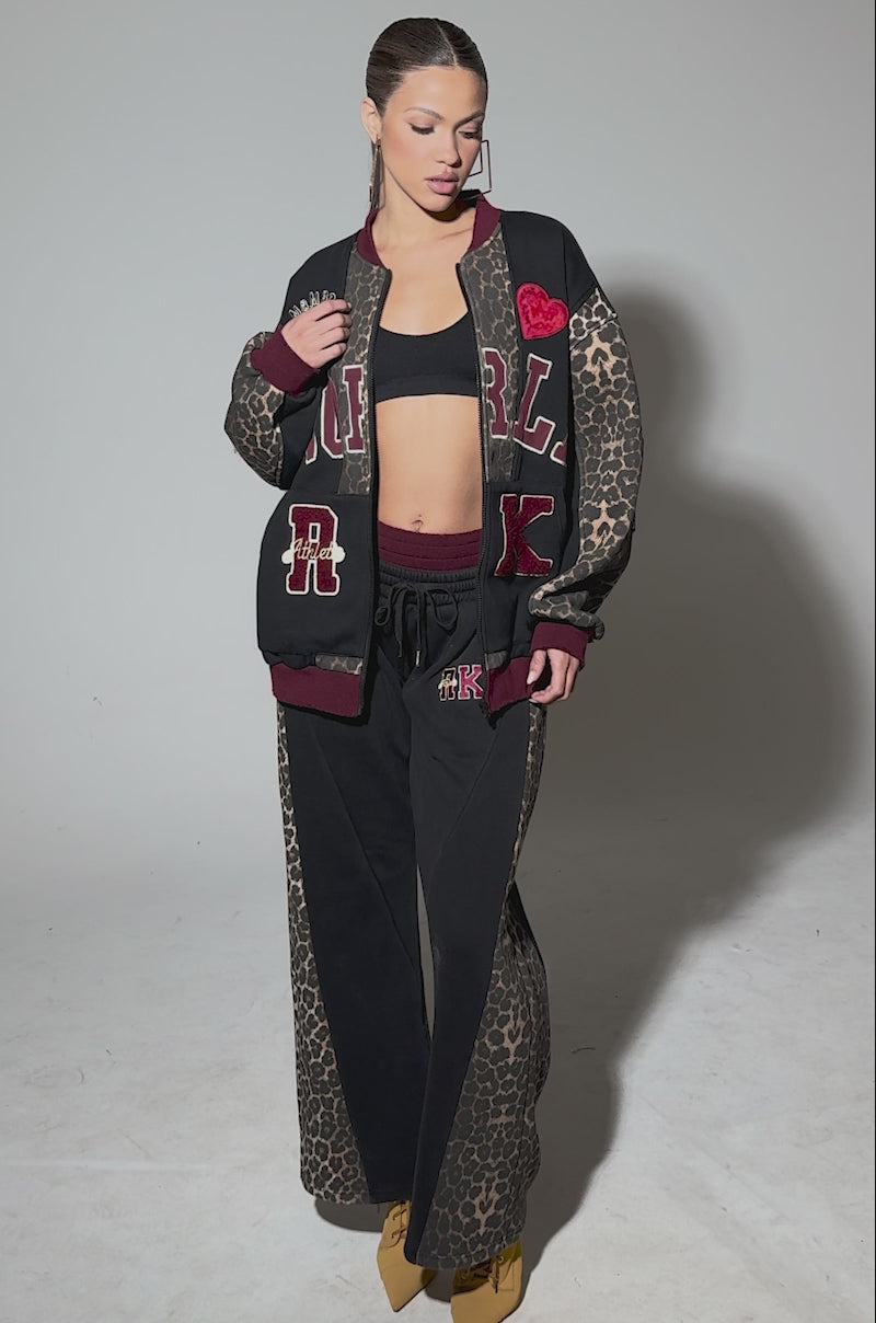 Video of woman modeling the WORLD HOLD ON ZIP UP SWEATSHIRT featuring a loose leopard print with bold patches—“NORL,” “RK,” a red heart, and a sports emblem. She wears hoop earrings and has her hair pulled back against a plain gray background.