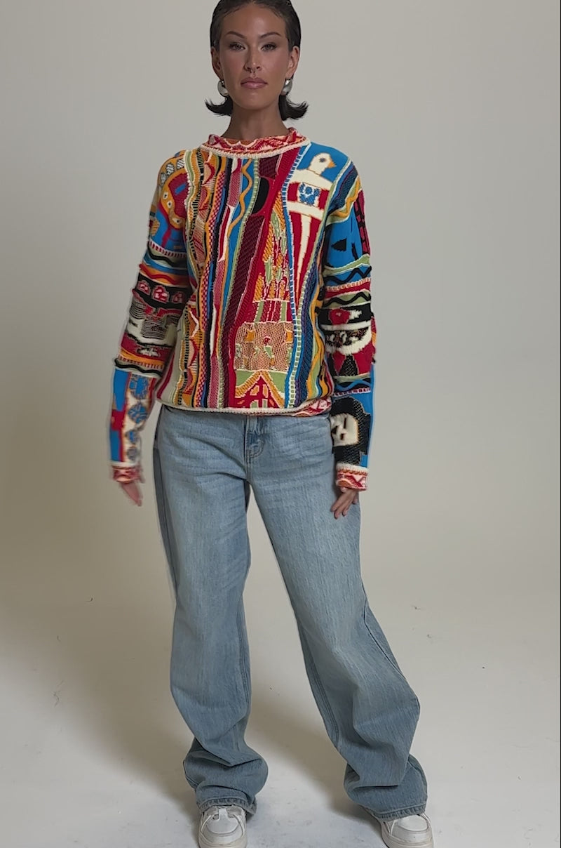 A video of a person with short dark hair and a neutral expression standing against a plain backdrop, wearing the RETRO OVERSIZED SWEATER IN MULTI, loose light blue jeans, and white sneakers.
