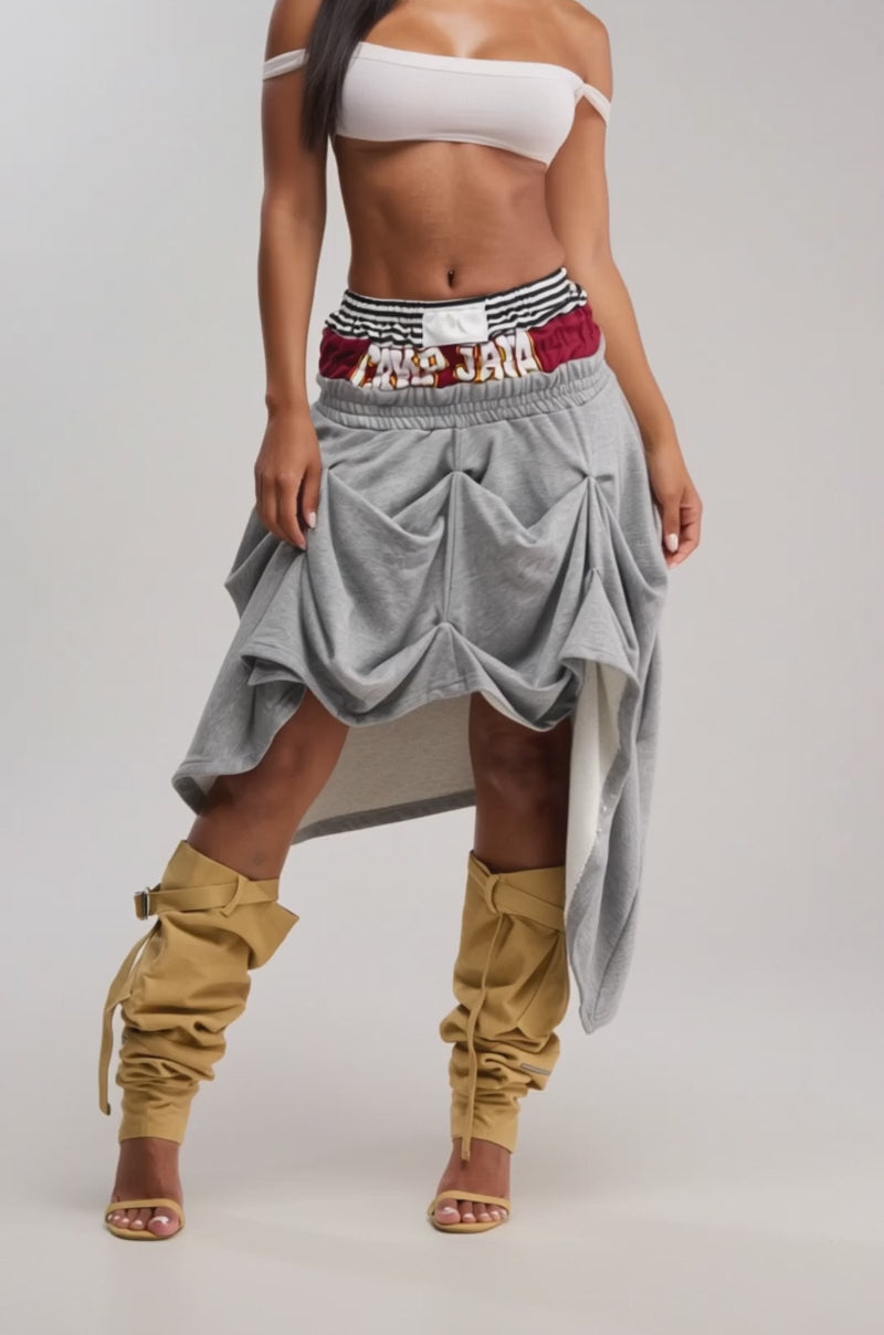 Video of woman modeling the NO REGRETS HERE MIDI SKIRT, a gray sporty ruched skirt with an asymmetrical hem and striped waistband, pairing it with tall mustard-yellow slouchy boots against a plain background.