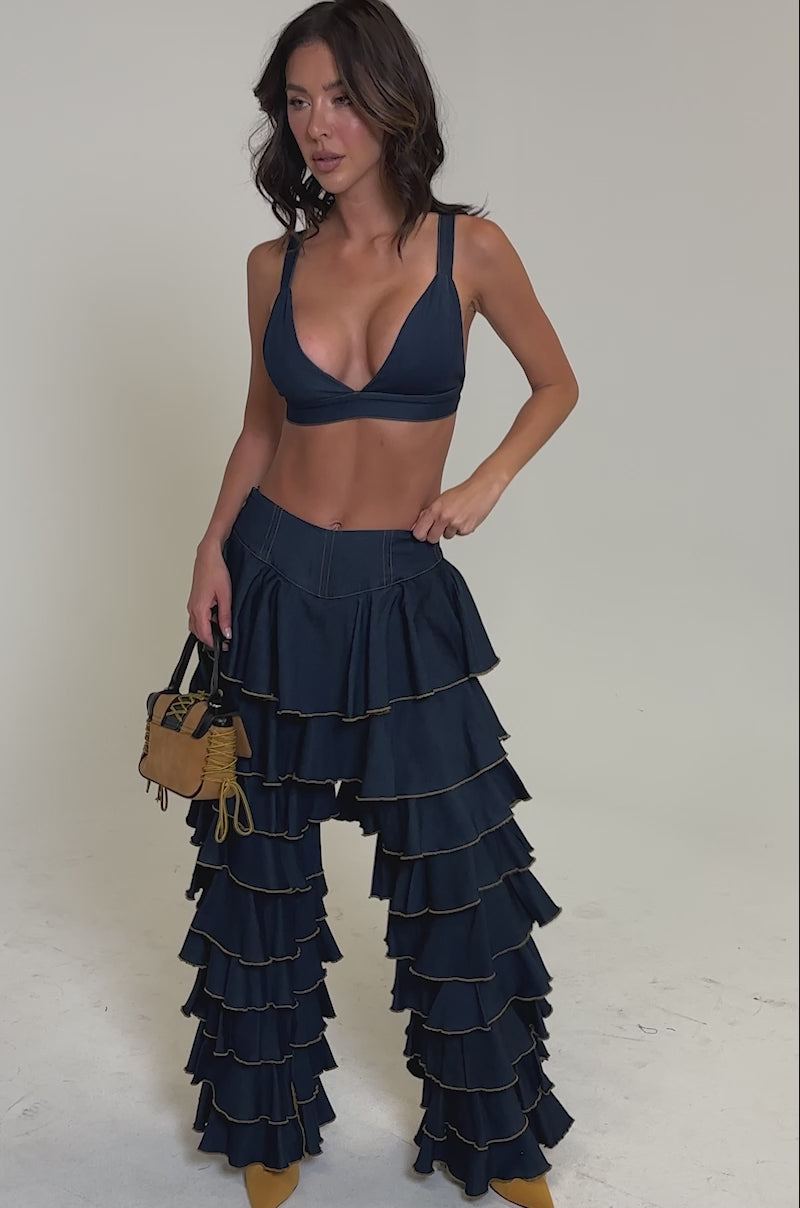 Video of woman modeling the SHAKE IT OUT DENIM RUFFLE JEANS and a deep blue bralette, accessorized with a brown fringe handbag and yellow heels, set against a plain light background.