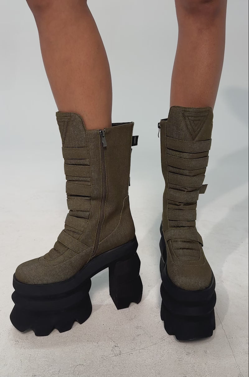 A video of a model wearing the AKIRA GOLD MENACING OLIVE BLOCK PLATFORM BOOTIE which features a chunky olive-green design with multiple horizontal straps around the shaft and a bold black multi-layered geometric platform sole.