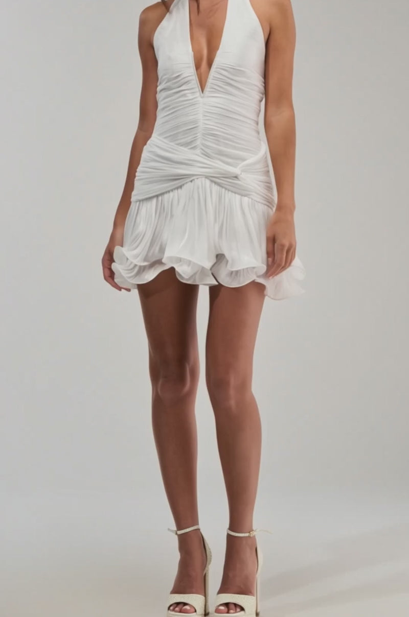 A video of a woman in the GOLDEN GLAM RUFFLE MINI DRESS IN WHITE, featuring a deep V-neck and ruffled skirt, poses against a light background.