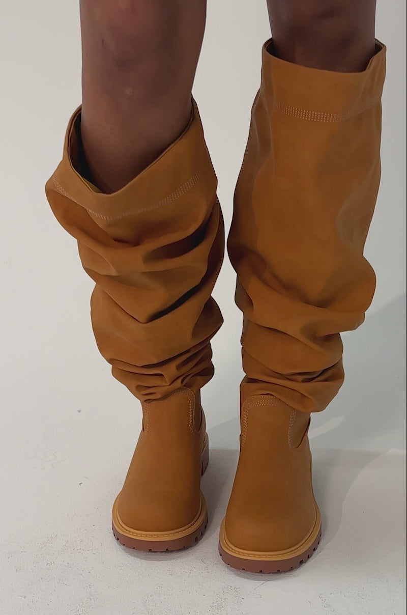 Video of woman modeling the AKIRA GOLD ARDIS Tan Ruched Flat Boots with rugged soles. The background is plain and light.