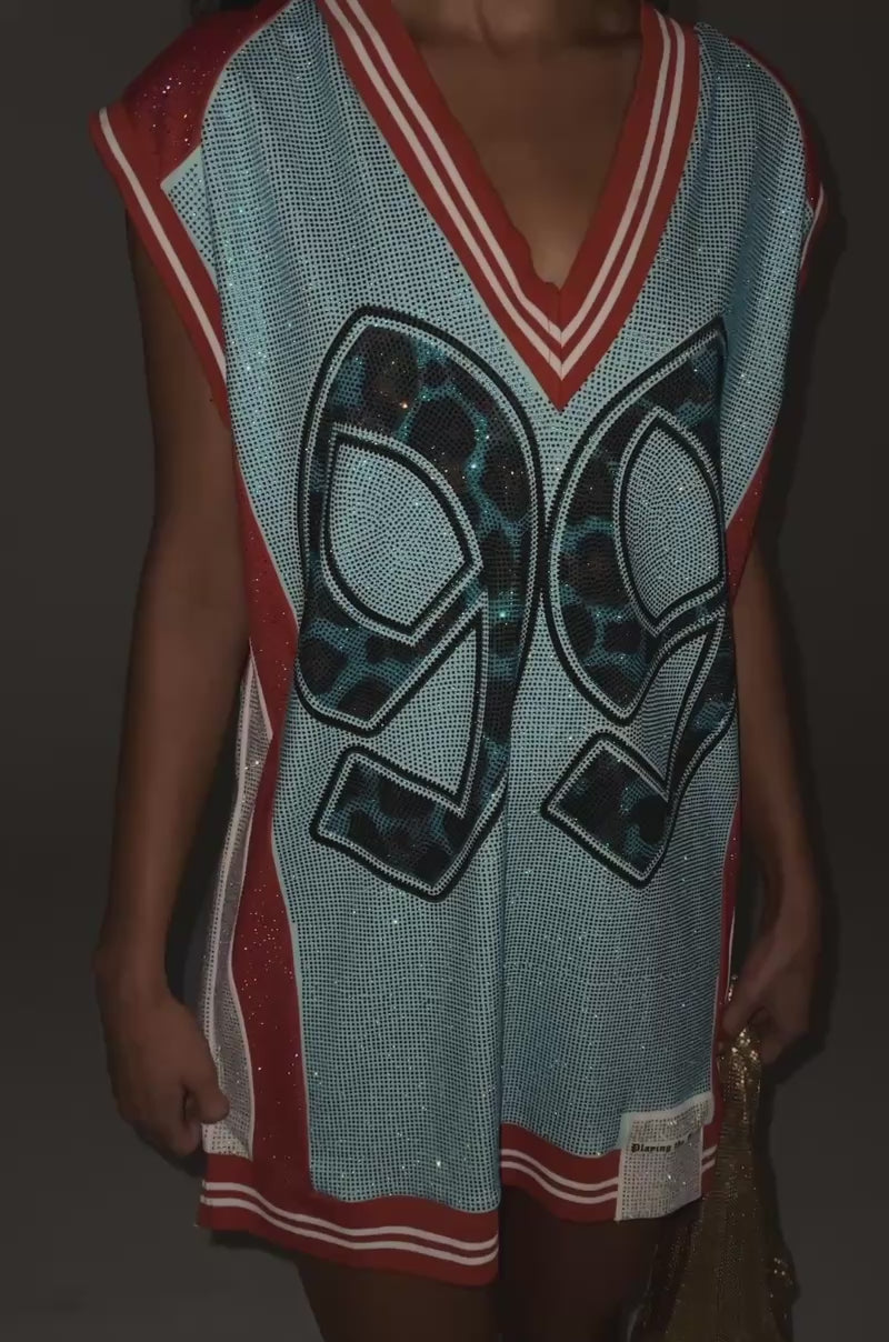 Video of woman modeling the RYAN COLORBLOCK SLEEVELESS JERSEY IN BLUE COMBO, a loose blue sport dress with red trim and a bold “99” on the front. She holds a gold bag and stands against a gray background.
